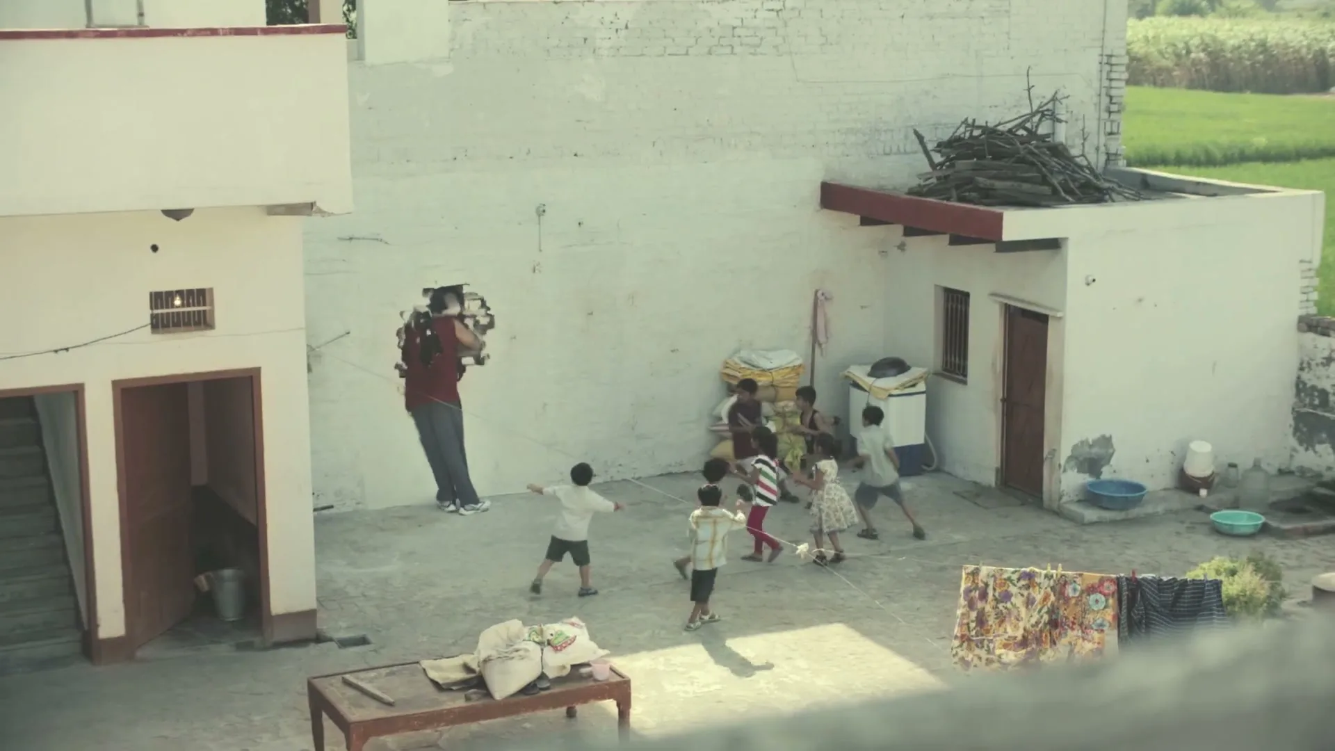 A giant man in a red shirt and blue pants plays with a group of children in a sunny, rural courtyard, accidentally breaking a wall.