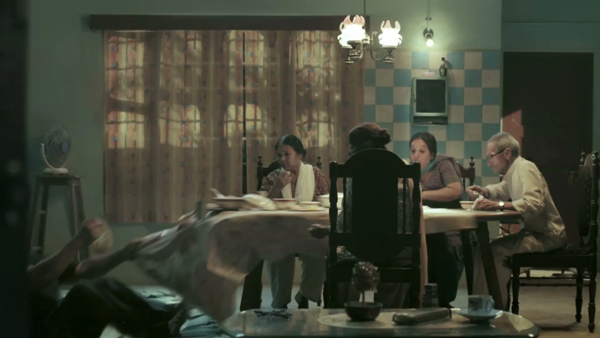 A very large man sits at a dining table with a family, then stands up and breaks the floor, causing the family to react in shock.