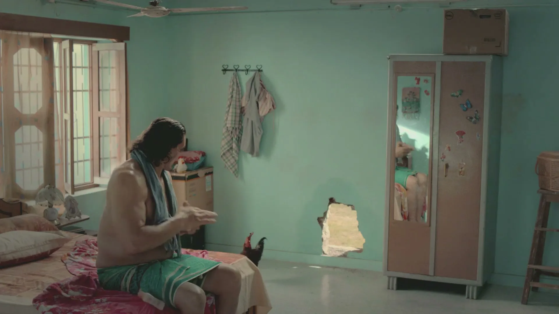 The Great Khali, a large man in a towel, sits on a bed in a bedroom, looking at a hole in the wall as a chicken walks through it.