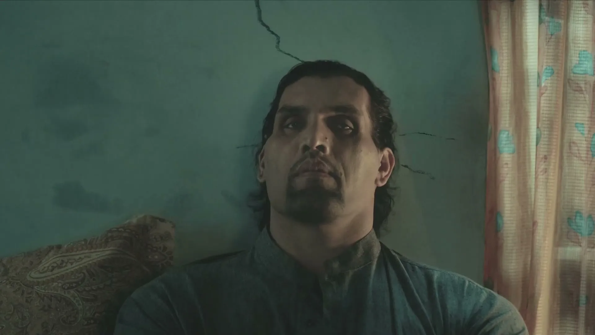 A man with dark hair and a beard sits against a cracked teal wall, looking up with a somber expression.
