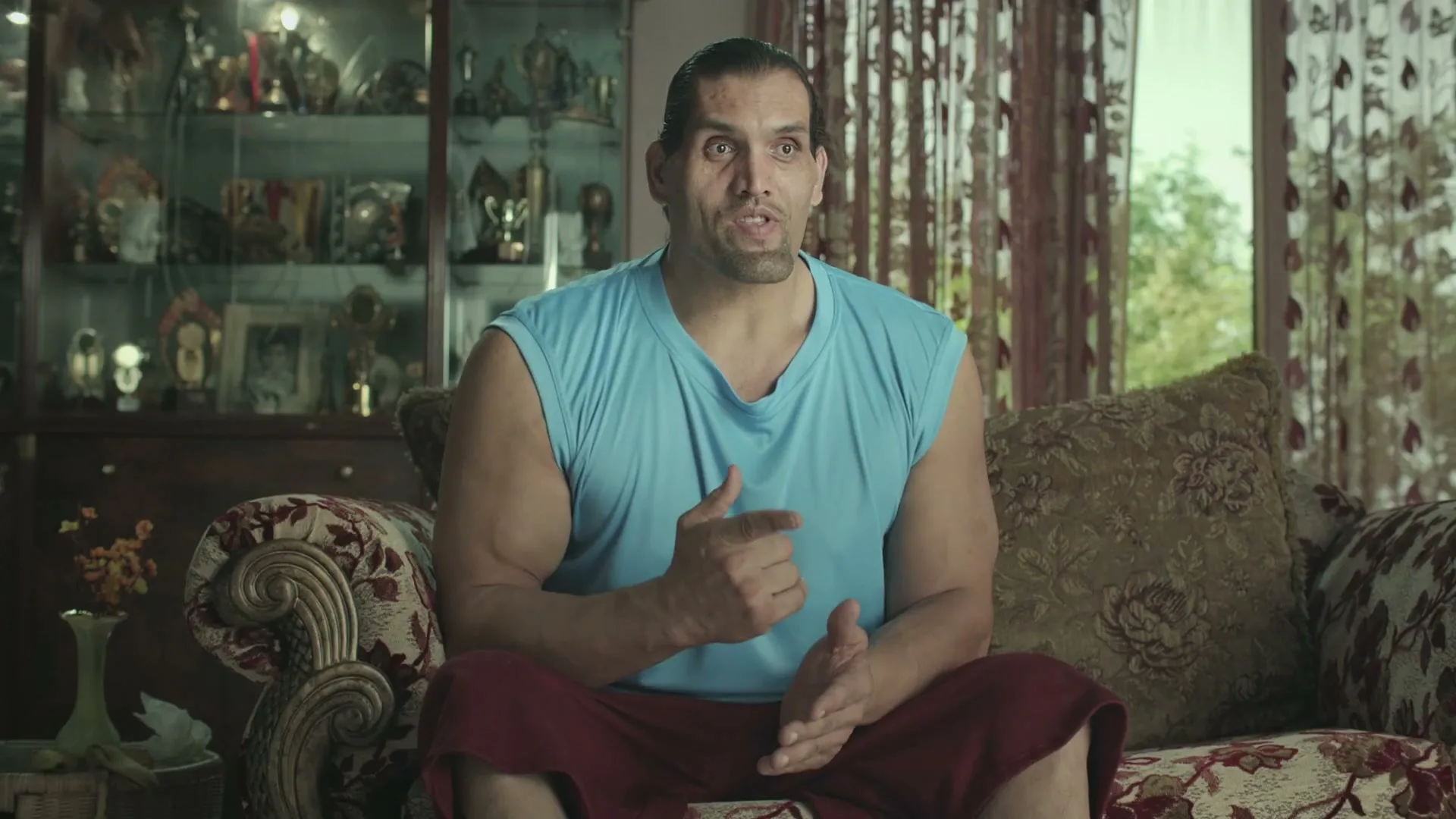 The Great Khali, a large muscular man, sits on a patterned sofa in a living room, talking and gesturing with his hands.