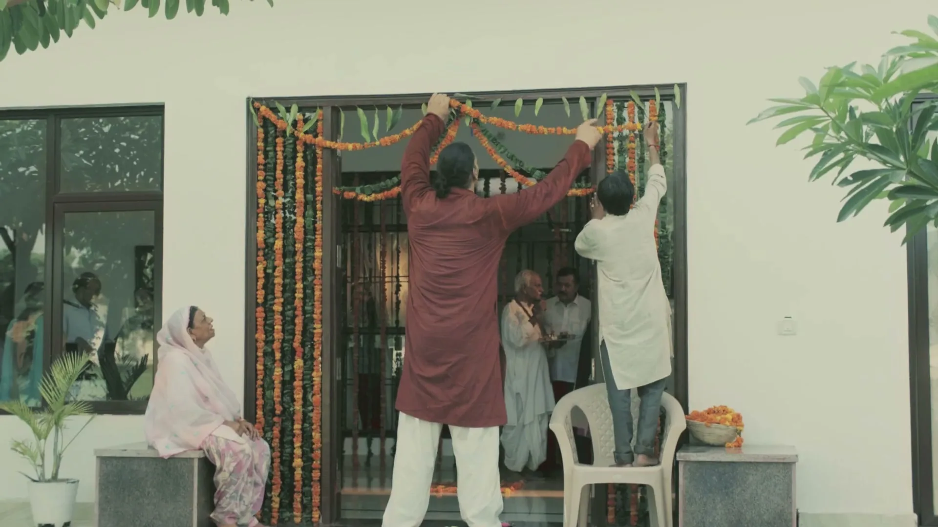 Two men decorate a doorway with marigold garlands while an older woman sits nearby and other people watch from inside a house.
