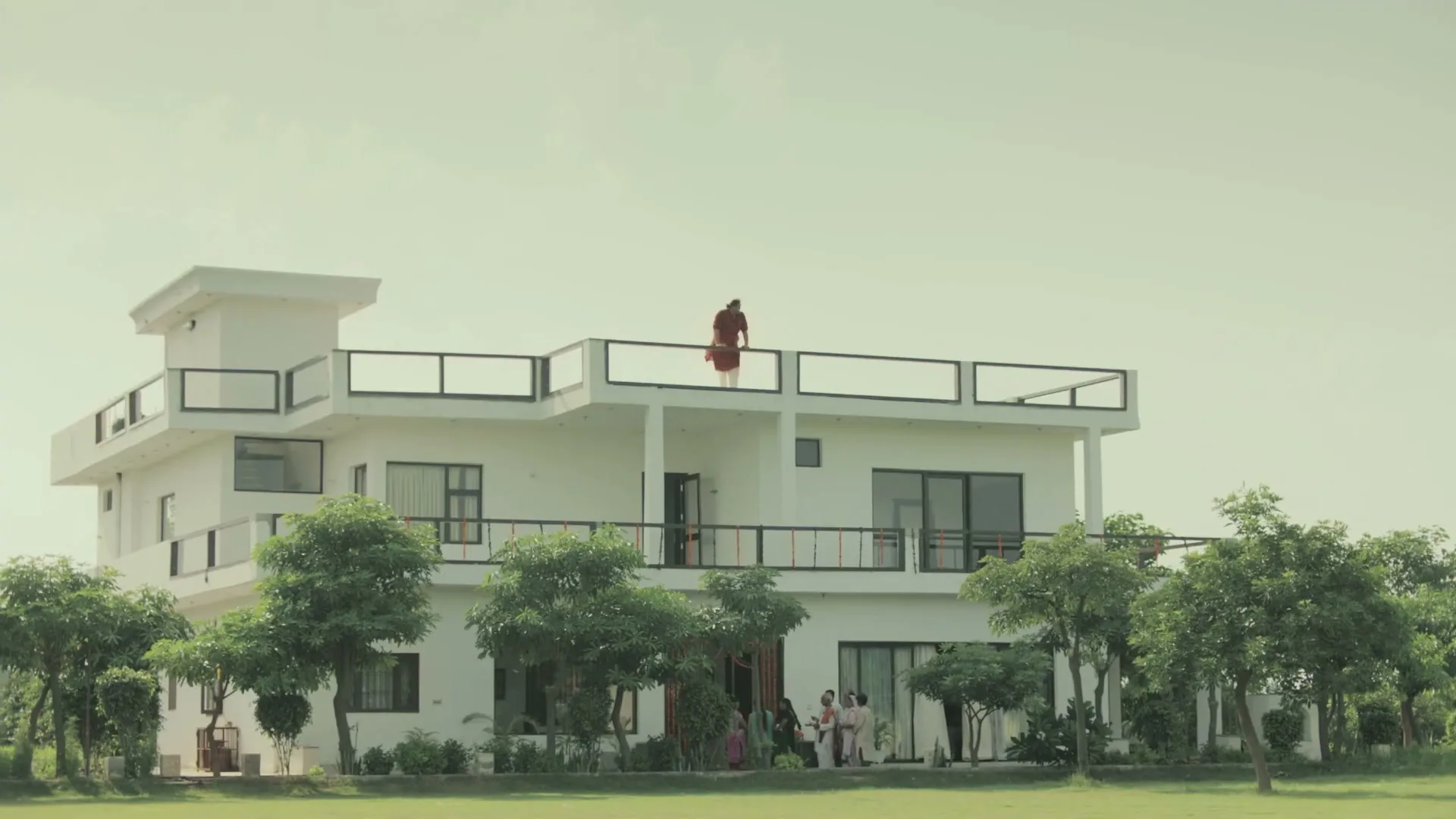 A man in a red shirt and white pants stands on the rooftop balcony of a large white house, looking down at a small group of people gathered below.