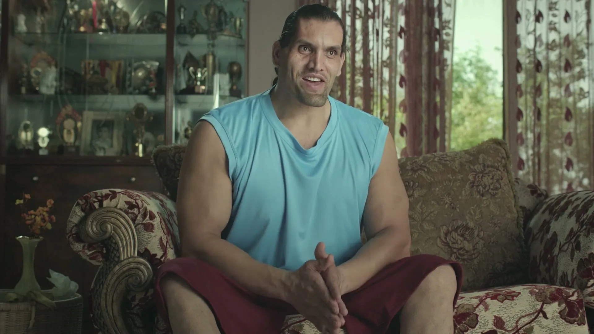 The Great Khali, a large male wrestler, sits on a patterned sofa in a living room, talking and gesturing with his hands.