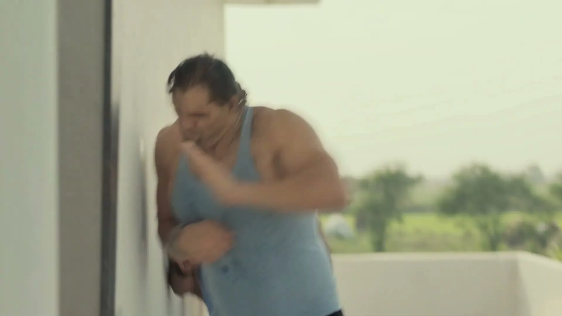 The Great Khali, a large male wrestler, in a blue tank top, pushes against a wall while two men watch him on a balcony.