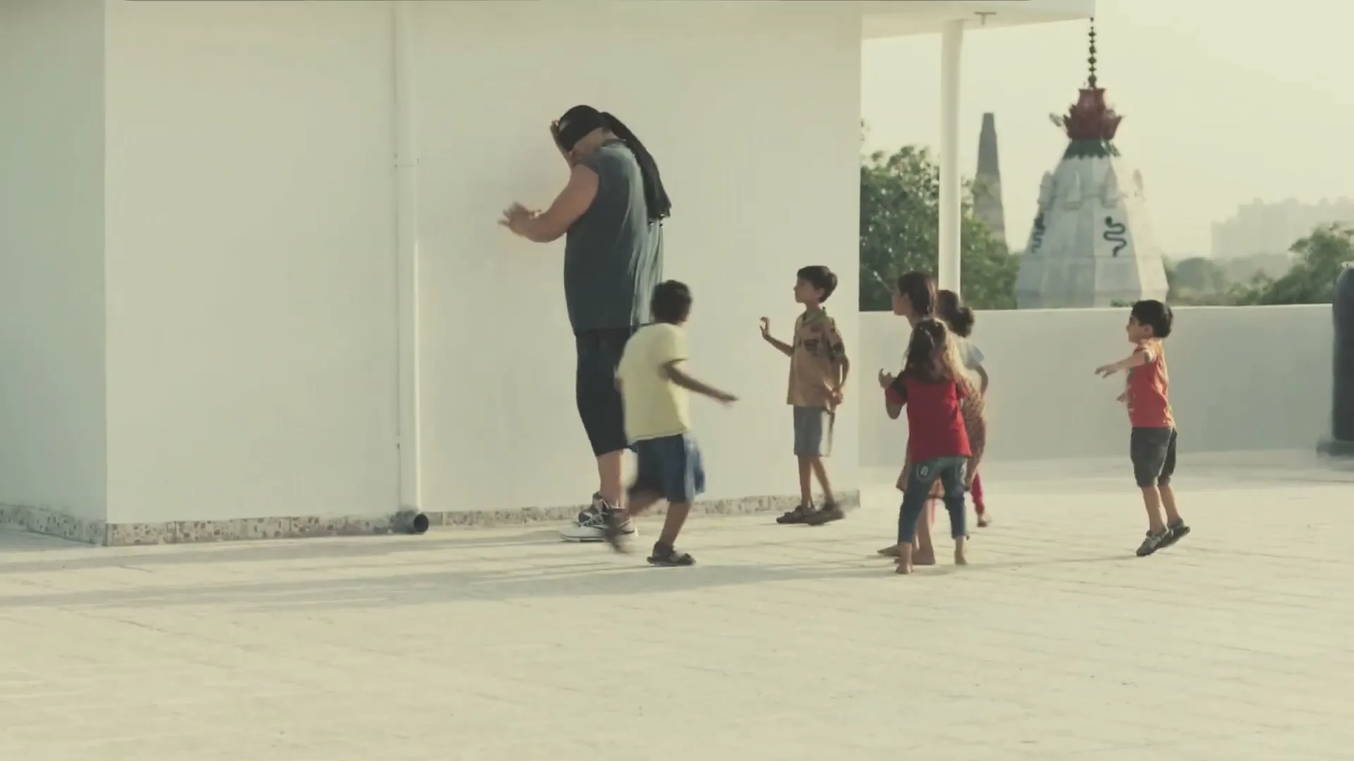 A giant man in a black tank top and shorts plays with a group of children on a rooftop, covering his eyes as they run around him.