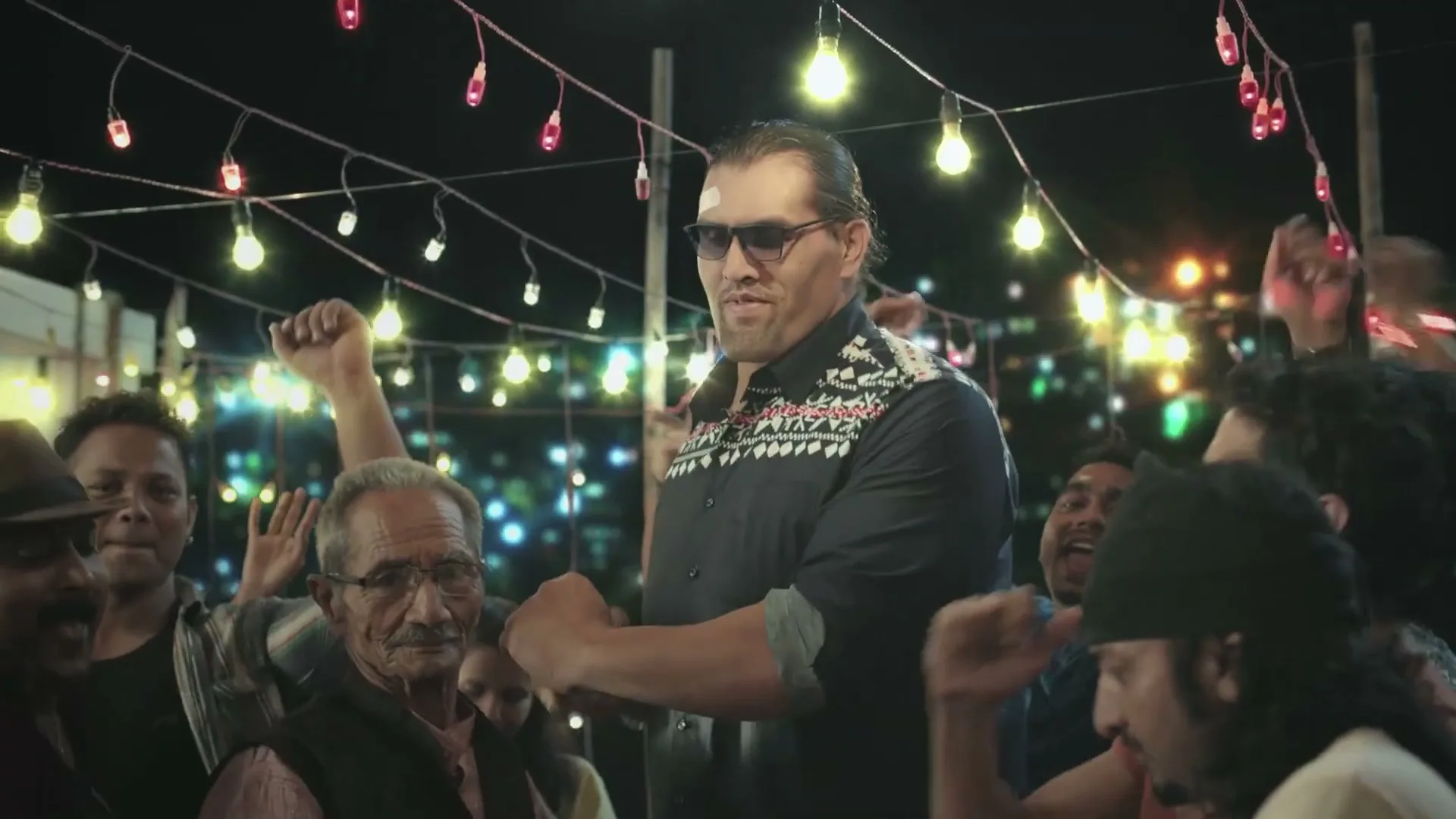 The Great Khali, a tall Indian man in sunglasses and a patterned shirt, dances with a crowd of people under string lights at night.