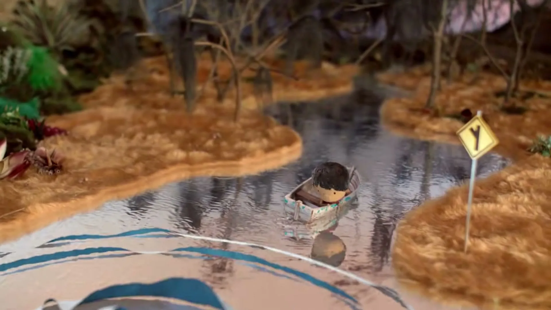 A small, dark-haired boy in a red shirt sits in a tiny boat, floating down a winding river in a stop-motion animated, fantastical landscape with a yellow road sign.