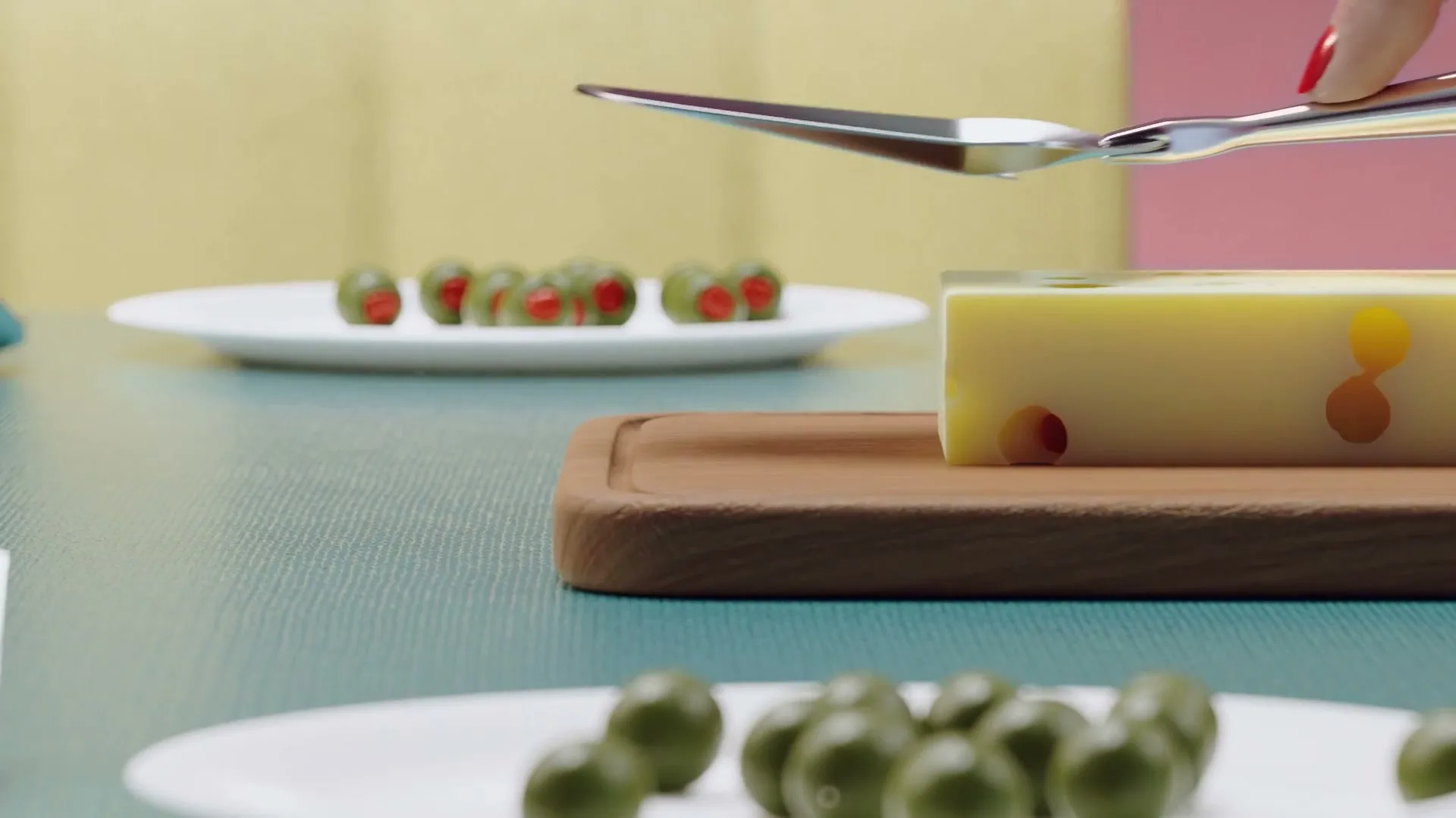 A woman's hand with red nail polish uses a cheese knife to cut a block of cheese with colorful holes on a wooden board, next to plates of olives on a table.
