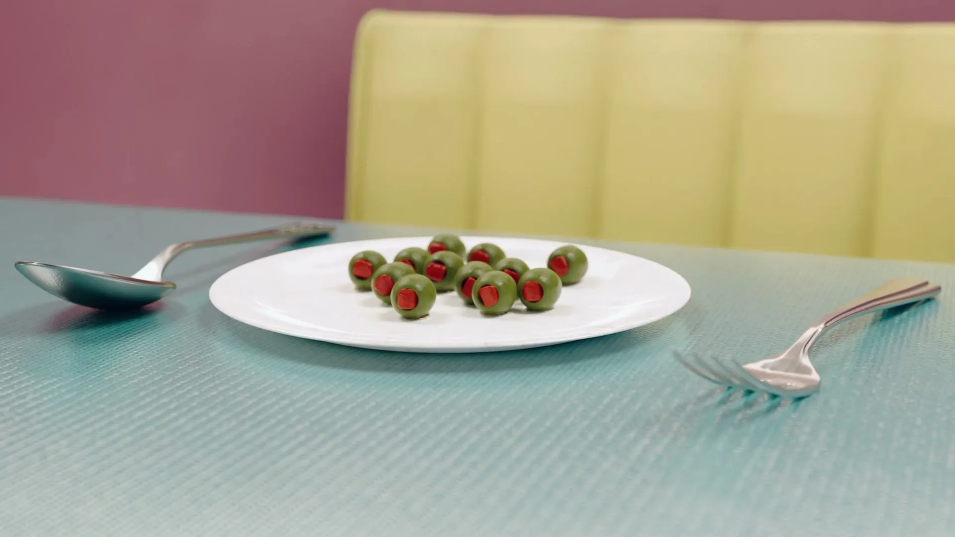 A white plate with green olives with red pimentos sits on a blue table with a spoon and fork, in a diner.