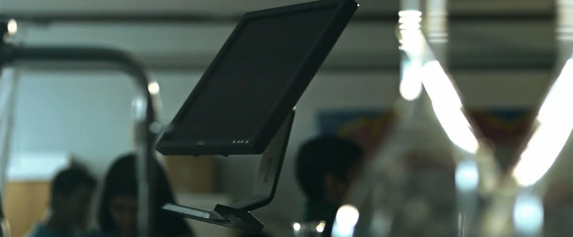 A close-up shot of a computer monitor on a desk, with blurred figures of people in the background, in an office setting.