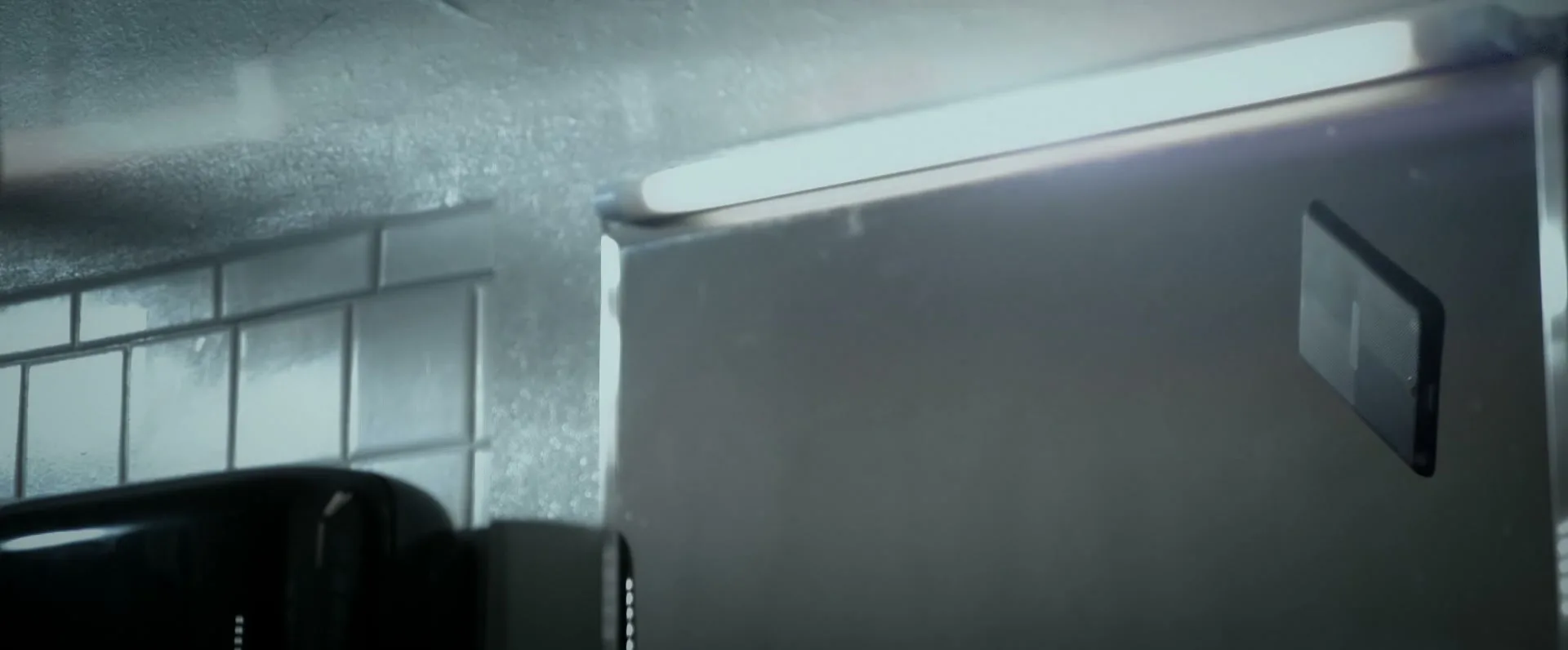 A black smartphone with a glowing screen is seen in a bathroom, reflecting light from a fluorescent bulb.
