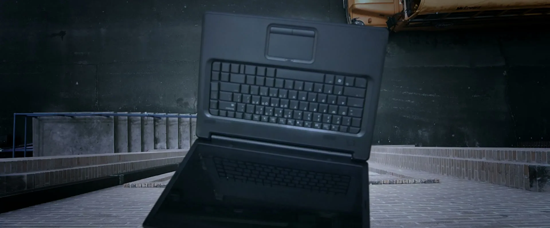 A black laptop falls through the air, seen from above, against a dark urban background with a yellow taxi cab visible.