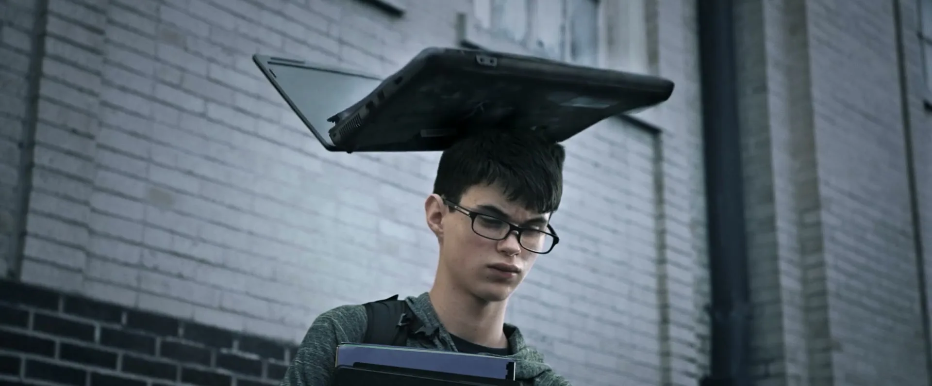 A young male student with glasses and a backpack stands in front of a brick wall as a laptop falls apart on his head in slow motion.