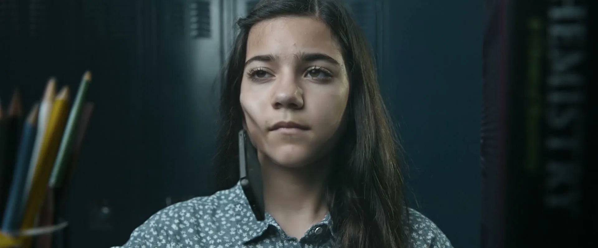 A young girl with long dark hair and a patterned shirt stands in a school hallway, looking forward with a serious expression, then closes her eyes.