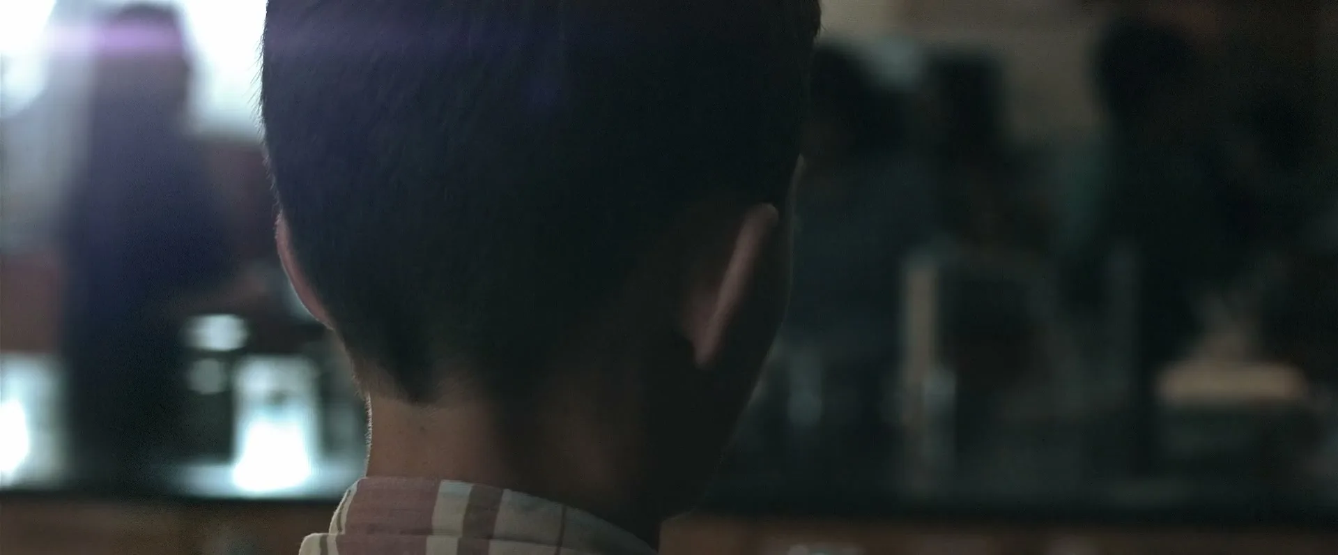 A young boy with dark hair and a plaid shirt stands with his back to the camera, looking towards a blurry background with bright lights.