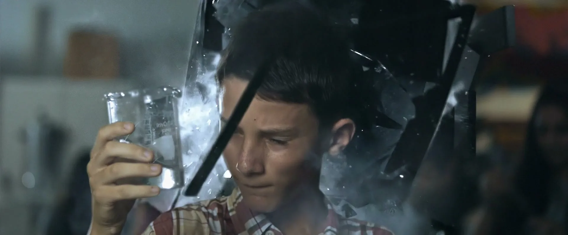 A young boy in a plaid shirt holds a beaker of water as glass shatters around him in a science classroom.