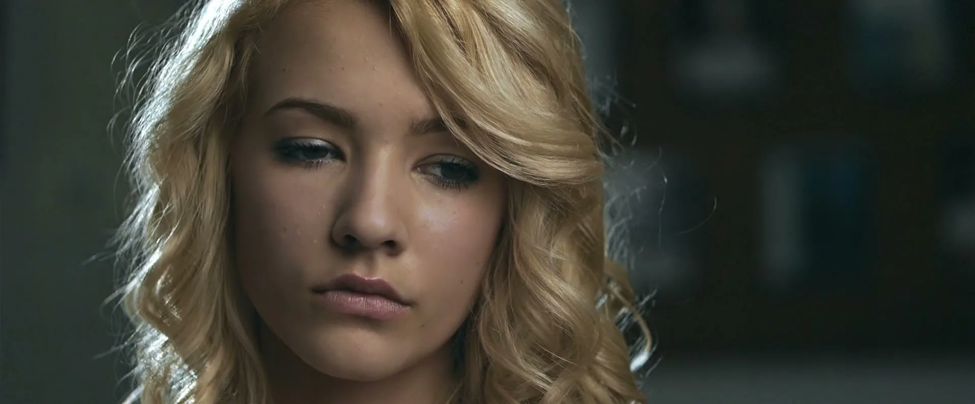 A young blonde woman with wavy hair looks down with a sad expression in a dimly lit interior setting.