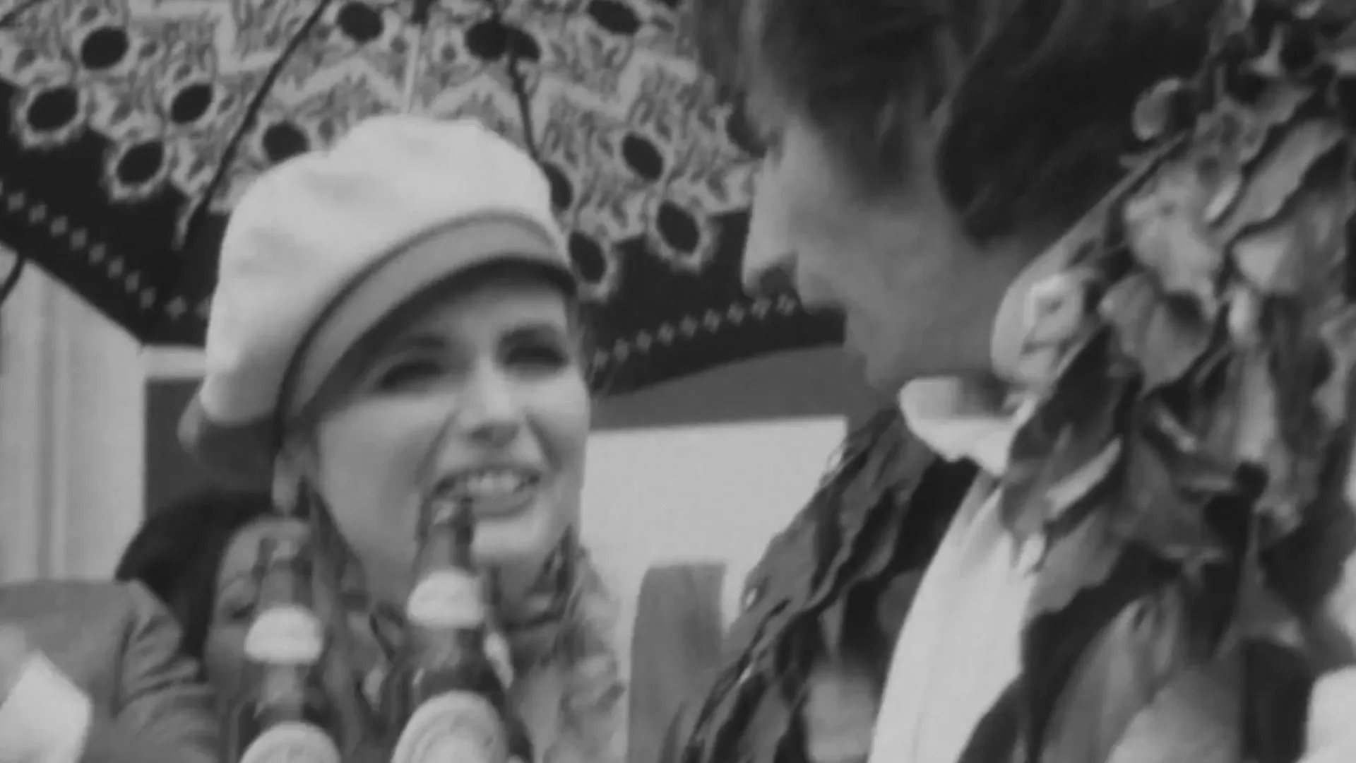 A black and white archival footage shot shows a young woman in a beret smiling and laughing as a man whispers into her ear, holding two Heineken bottles on a tray.