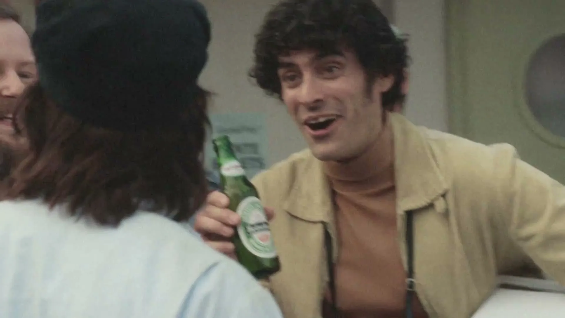A man with curly hair in a tan jacket talks to another person, holding a green beer bottle at a lively outdoor gathering.