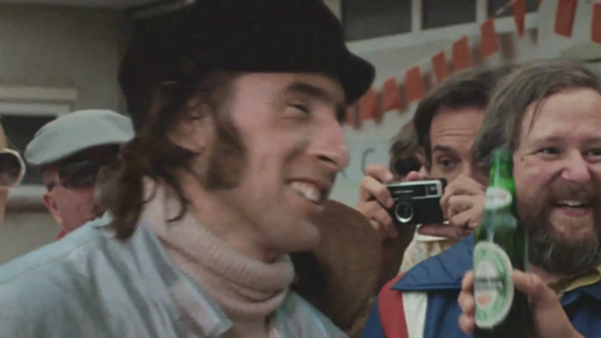 A man in a black hat and light blue jacket smiles while surrounded by a small group of people, one holding a camera and another a Heineken bottle, at an outdoor event.