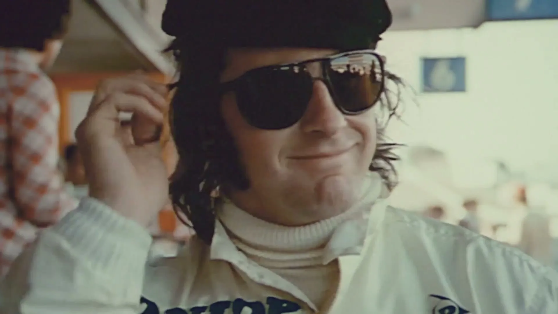 A man in a white racing suit, black cap, and sunglasses smiles while adjusting his cap in a pit lane.