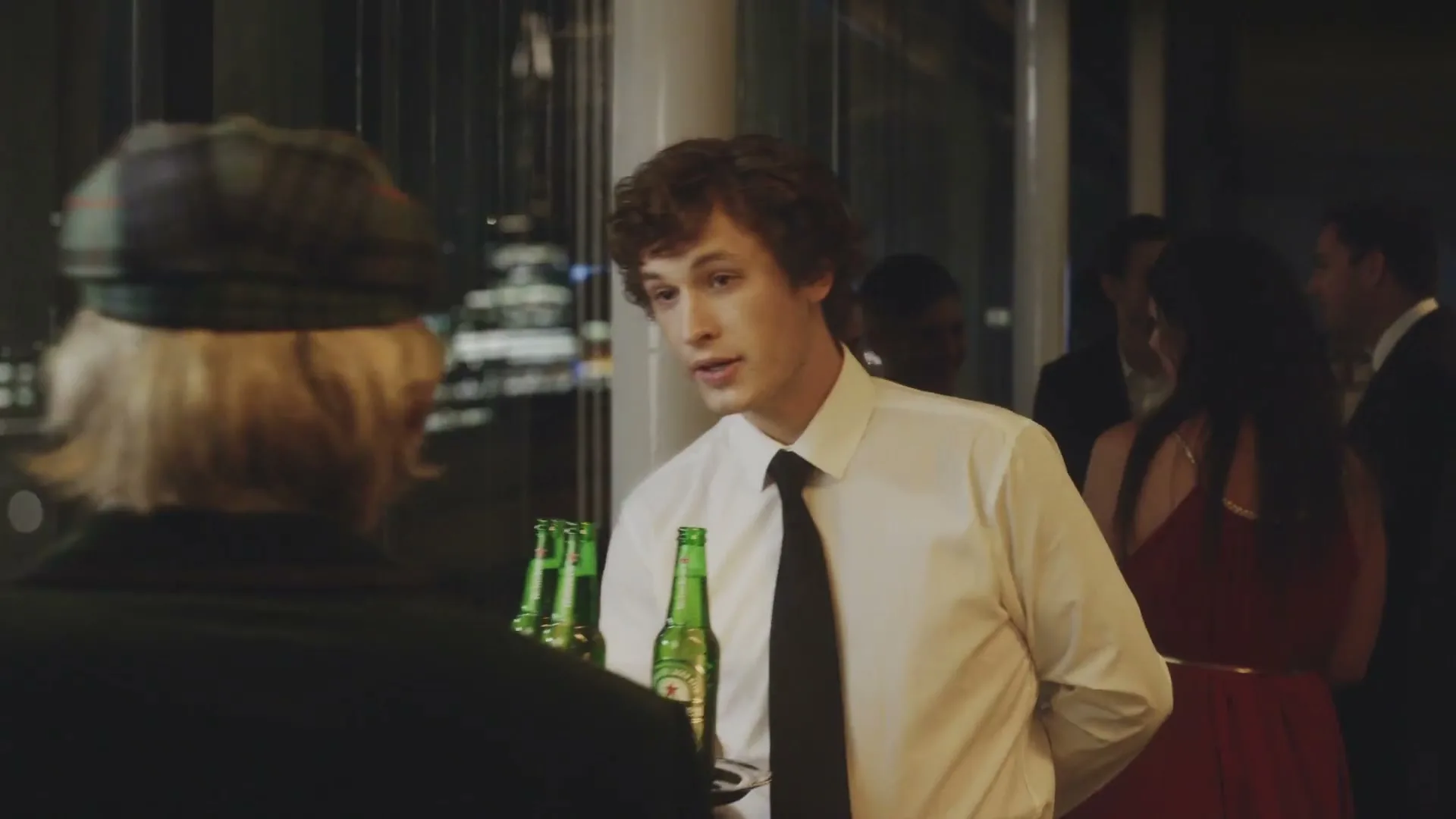 A young male waiter in a white shirt and black tie holds a tray of Heineken bottles while talking to an older person at a party.