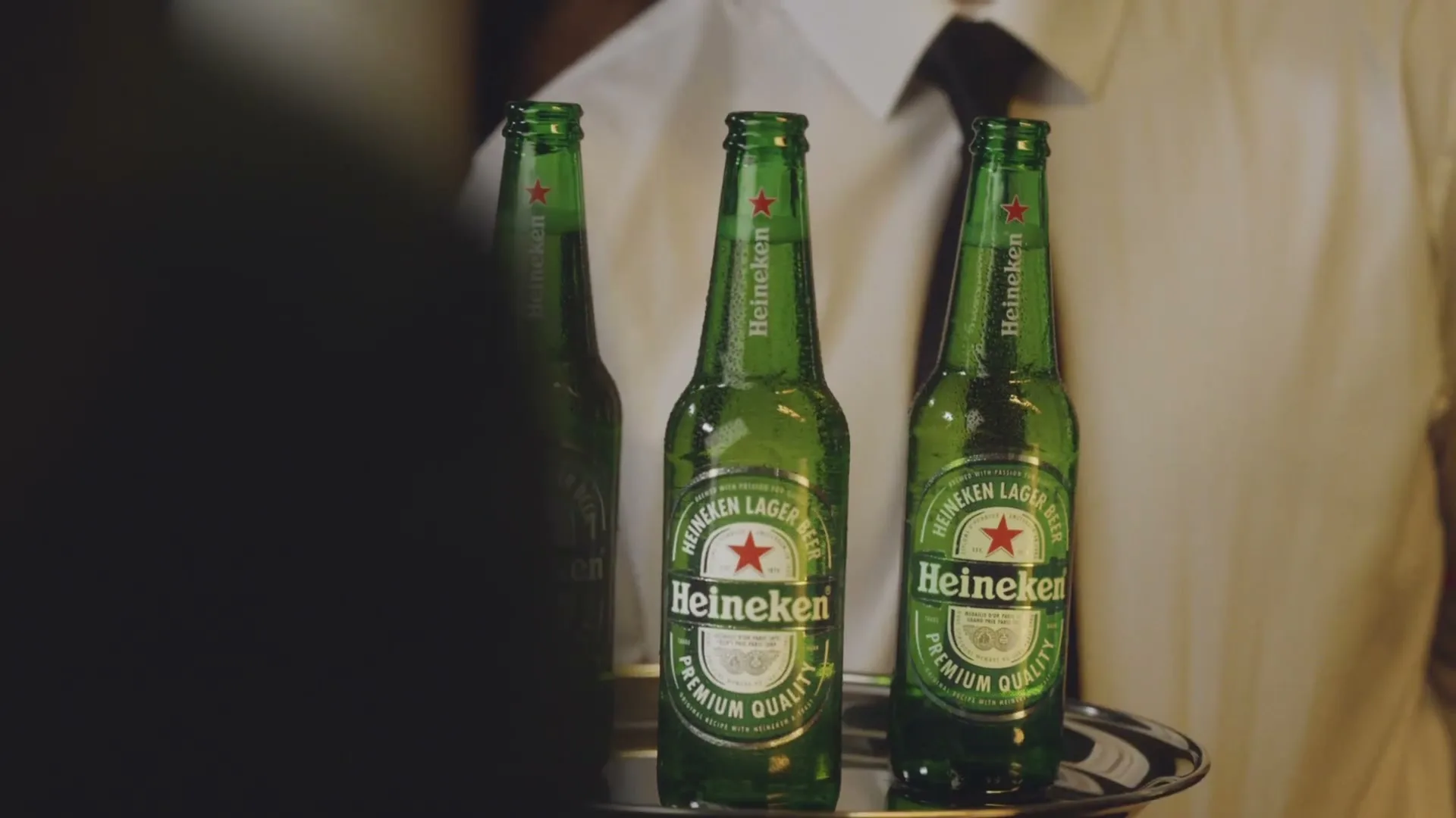 Three cold Heineken beer bottles are presented on a silver tray by a waiter in a restaurant or bar setting.