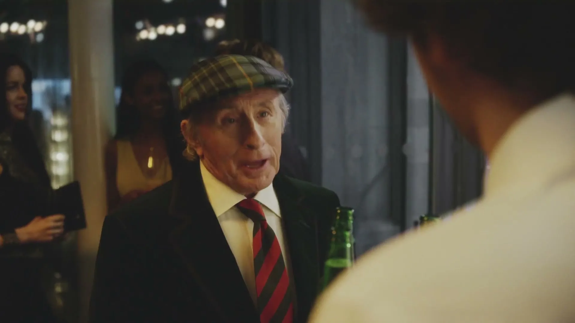 An older man in a plaid cap and suit talks to another person at a bar, declining a drink.