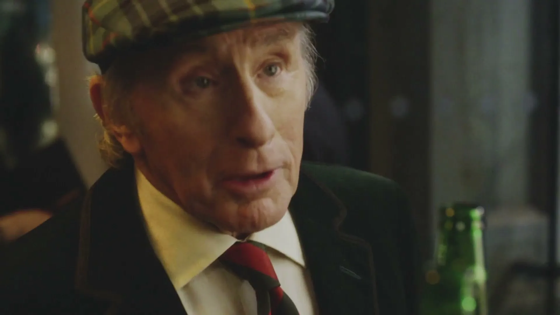 An older man in a plaid cap and suit jacket talks to someone off-screen in a dimly lit bar, then winks and adjusts his cap.