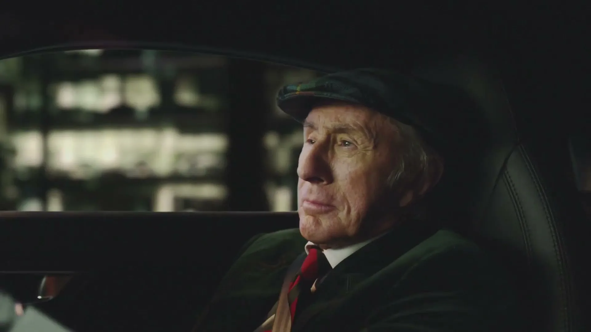 An older man in a flat cap and green jacket sits in the driver's seat of a car at night, looking out the window.