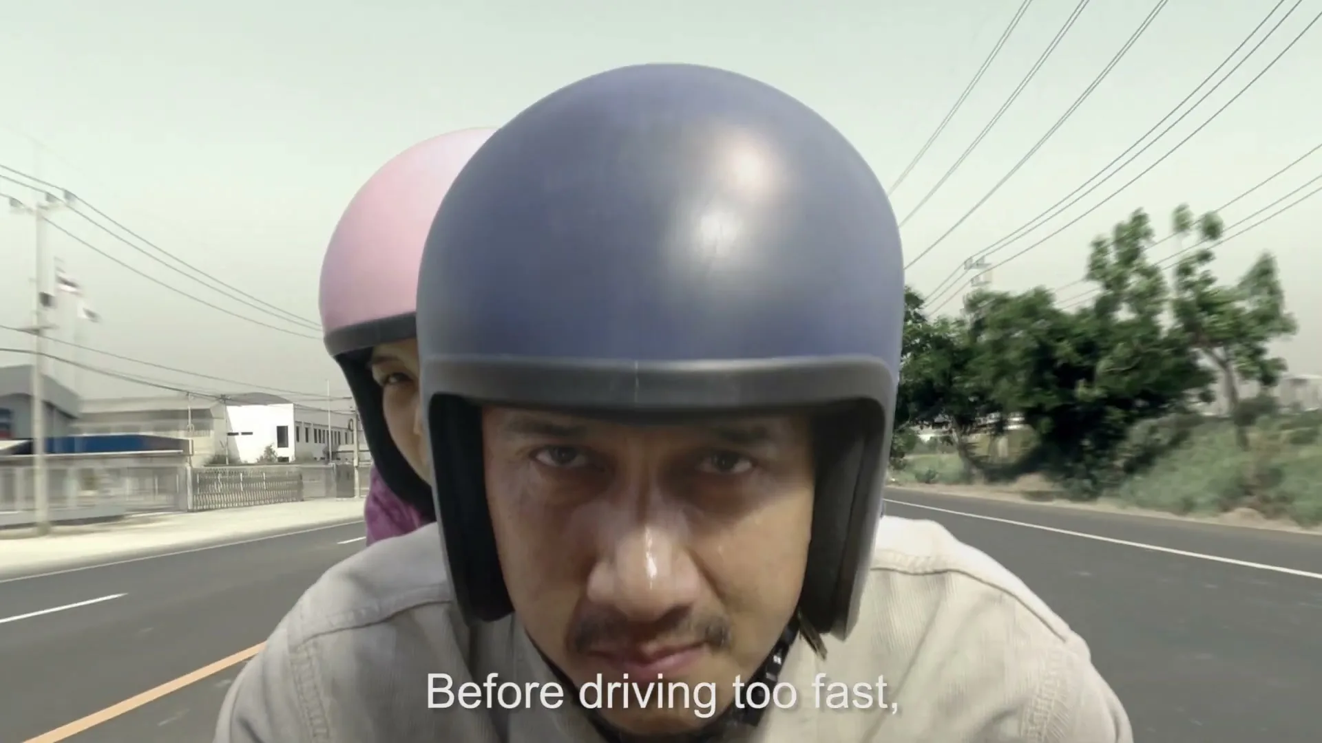 A man in a blue helmet drives a motorcycle with a woman in a pink helmet behind him, looking intensely at the camera on a road during the day.