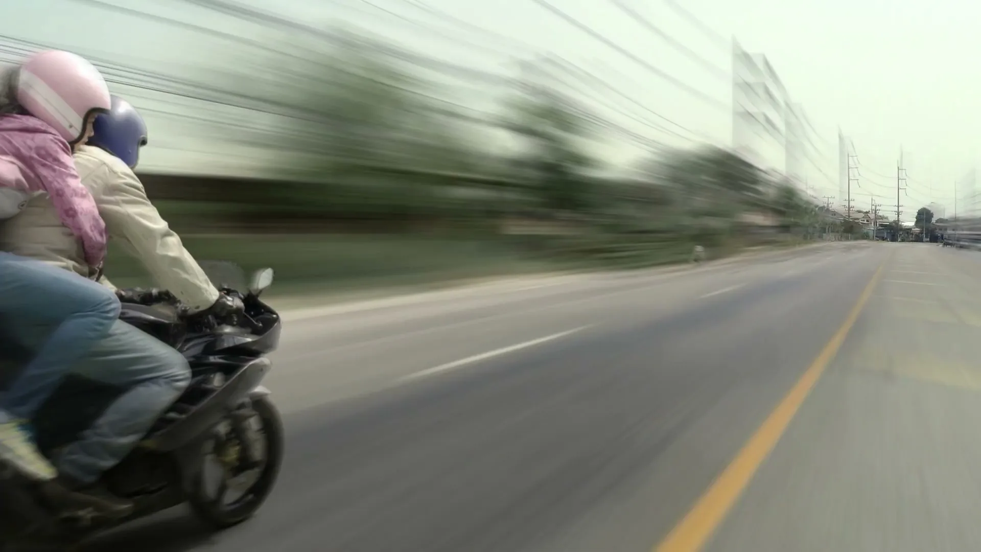 A man and a woman on a motorcycle speed down a road, with motion blur indicating high speed, on a bright day.