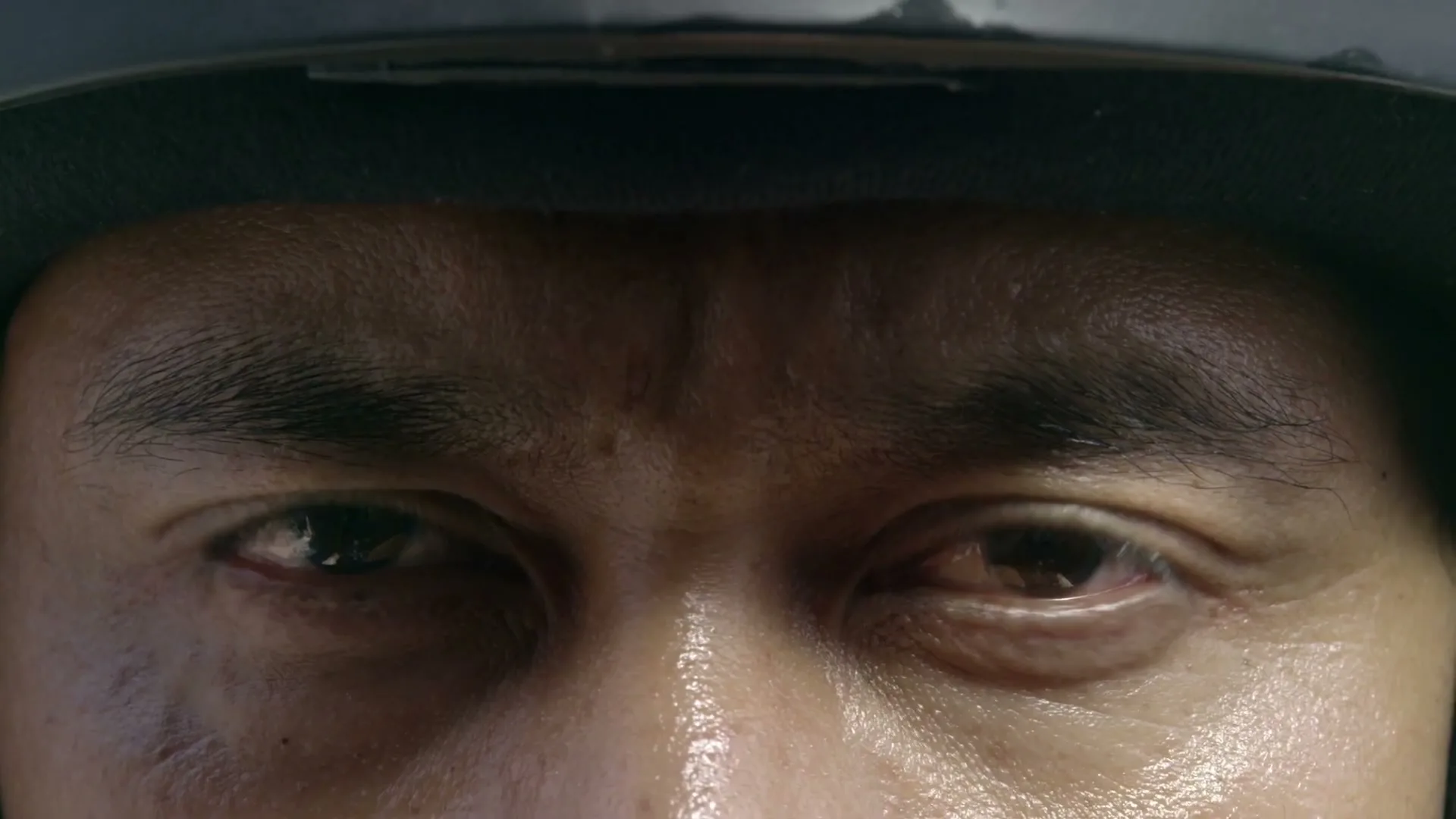 A man's eyes, wearing a helmet, widen in shock and fear, as if witnessing something horrific.
