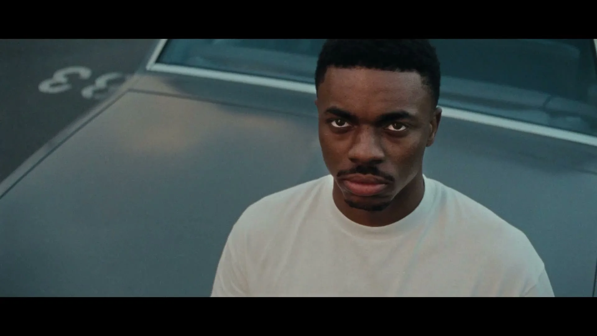 A young Black man with a serious expression looks directly at the camera, standing in front of a grey car in an outdoor setting.
