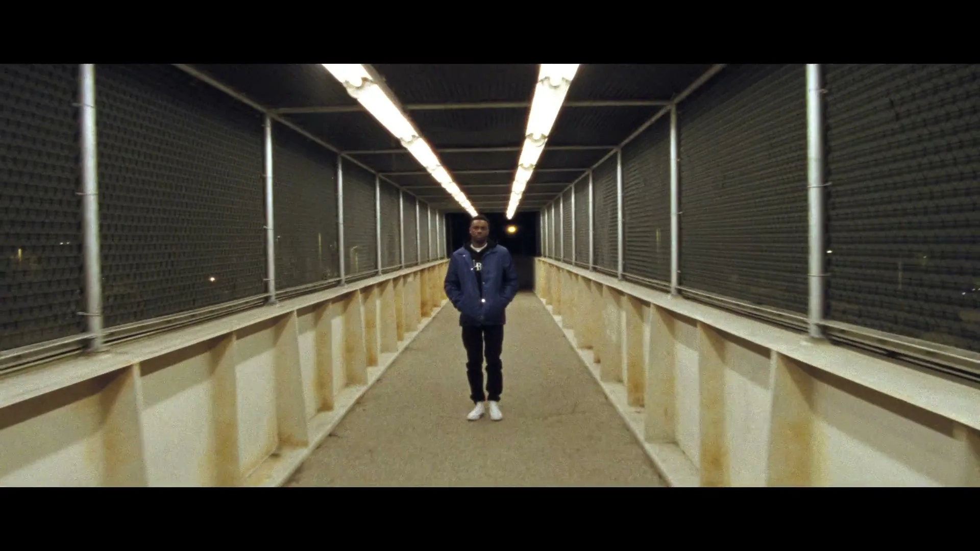 A young man in a blue jacket and white sneakers stands still in a brightly lit pedestrian bridge at night.