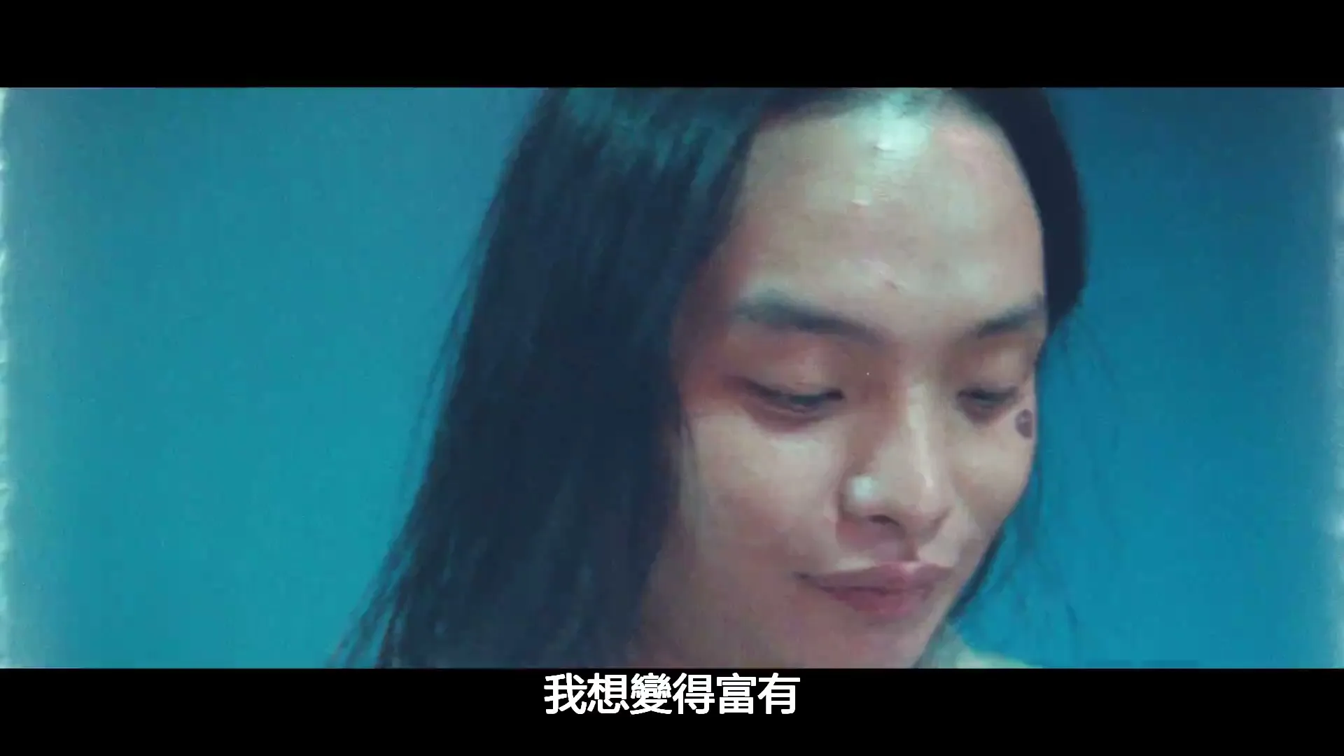 A young woman with long dark hair and a tattoo on her cheek looks down, then the screen flashes to white, in a studio.