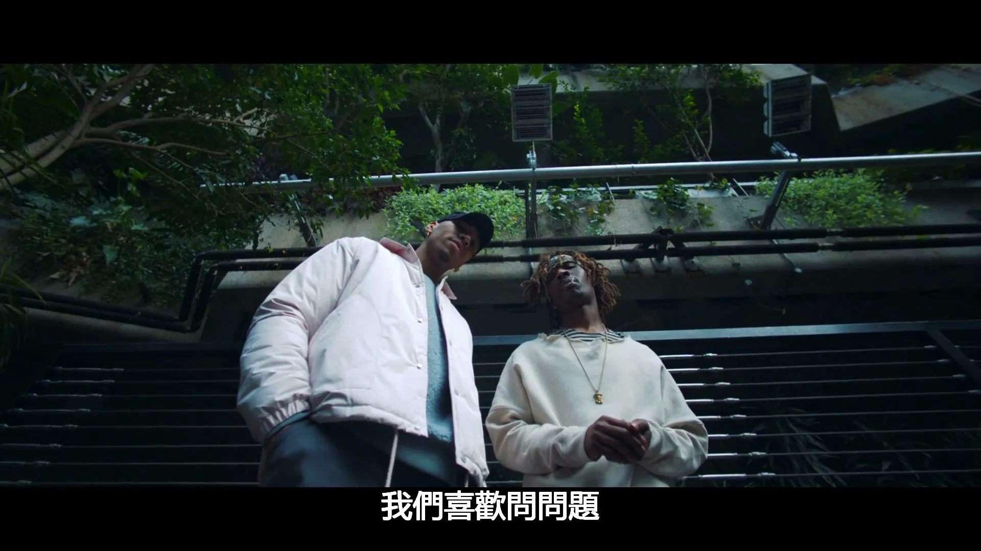 Two young men, one in a white jacket and the other in a cream sweatshirt, stand looking up in an urban setting with greenery.