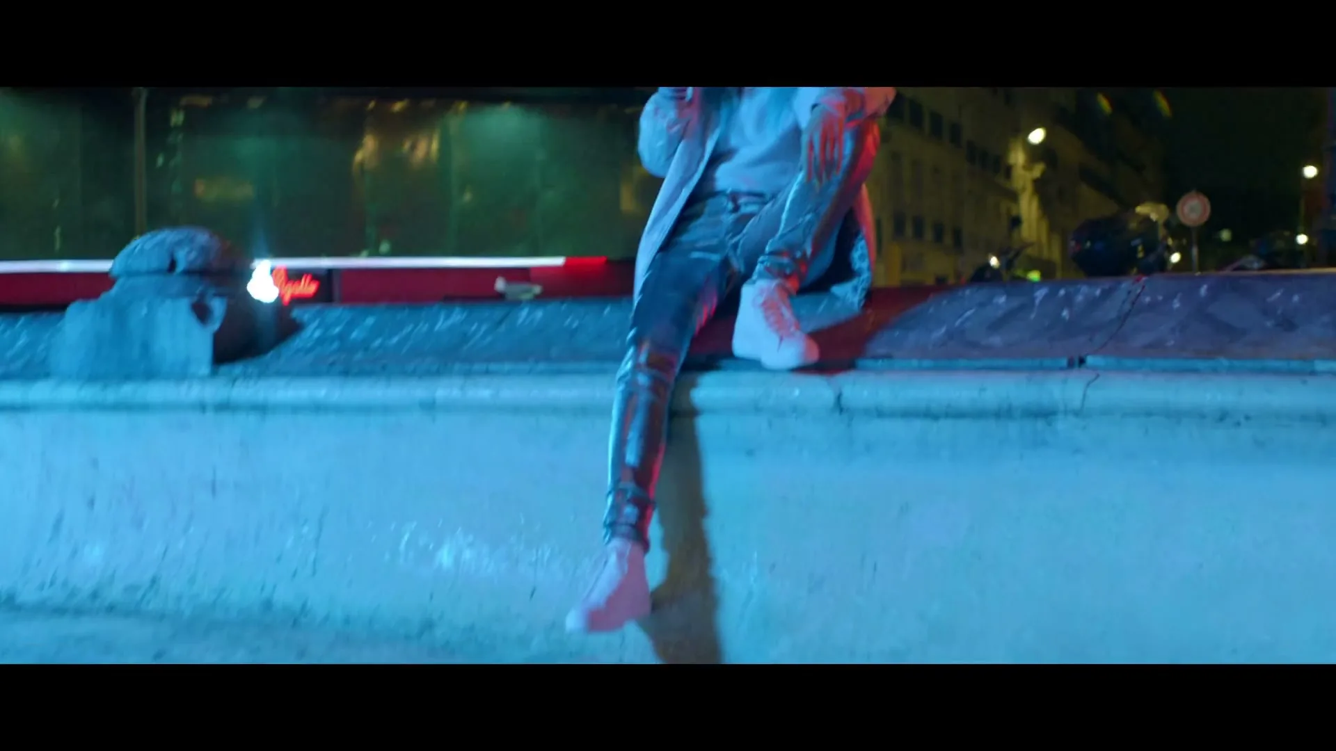 A young woman with blonde hair, wearing a white coat and shiny pants, sits on a ledge at night, drinking from a cup, with a bus in the background.