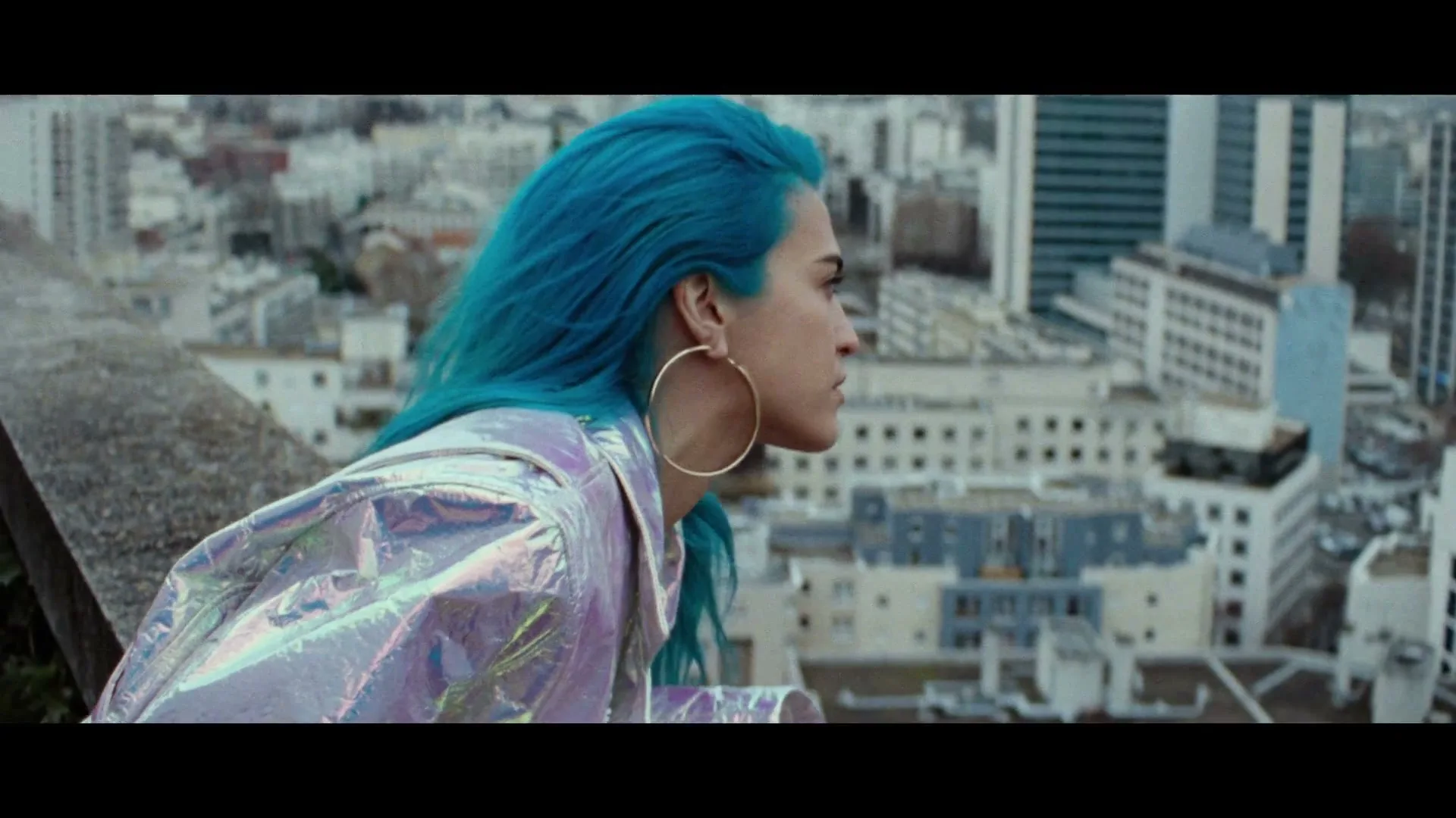 A young woman with bright blue hair and a shiny jacket looks out over a city from a rooftop.
