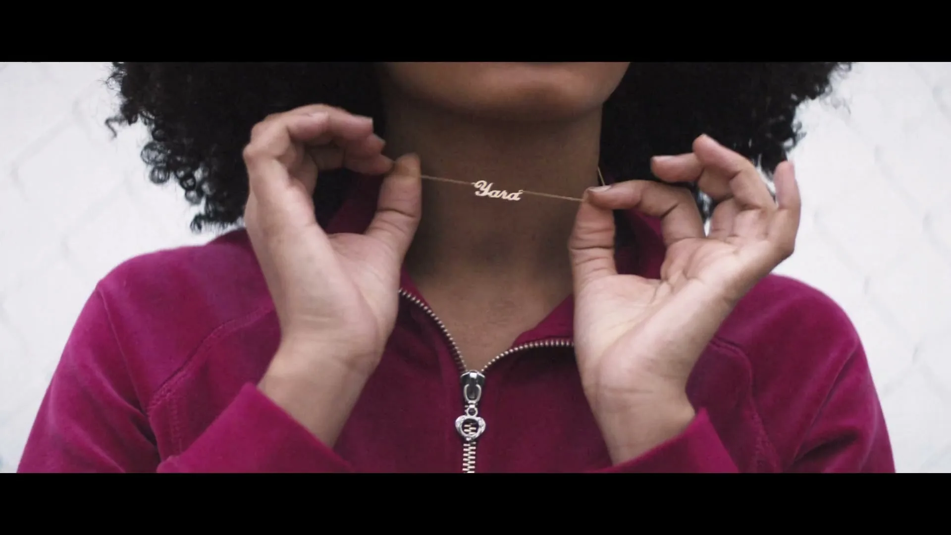 A young woman with curly hair wears a pink velour tracksuit and holds a gold necklace with the name "Yara" on it, in an exterior setting.