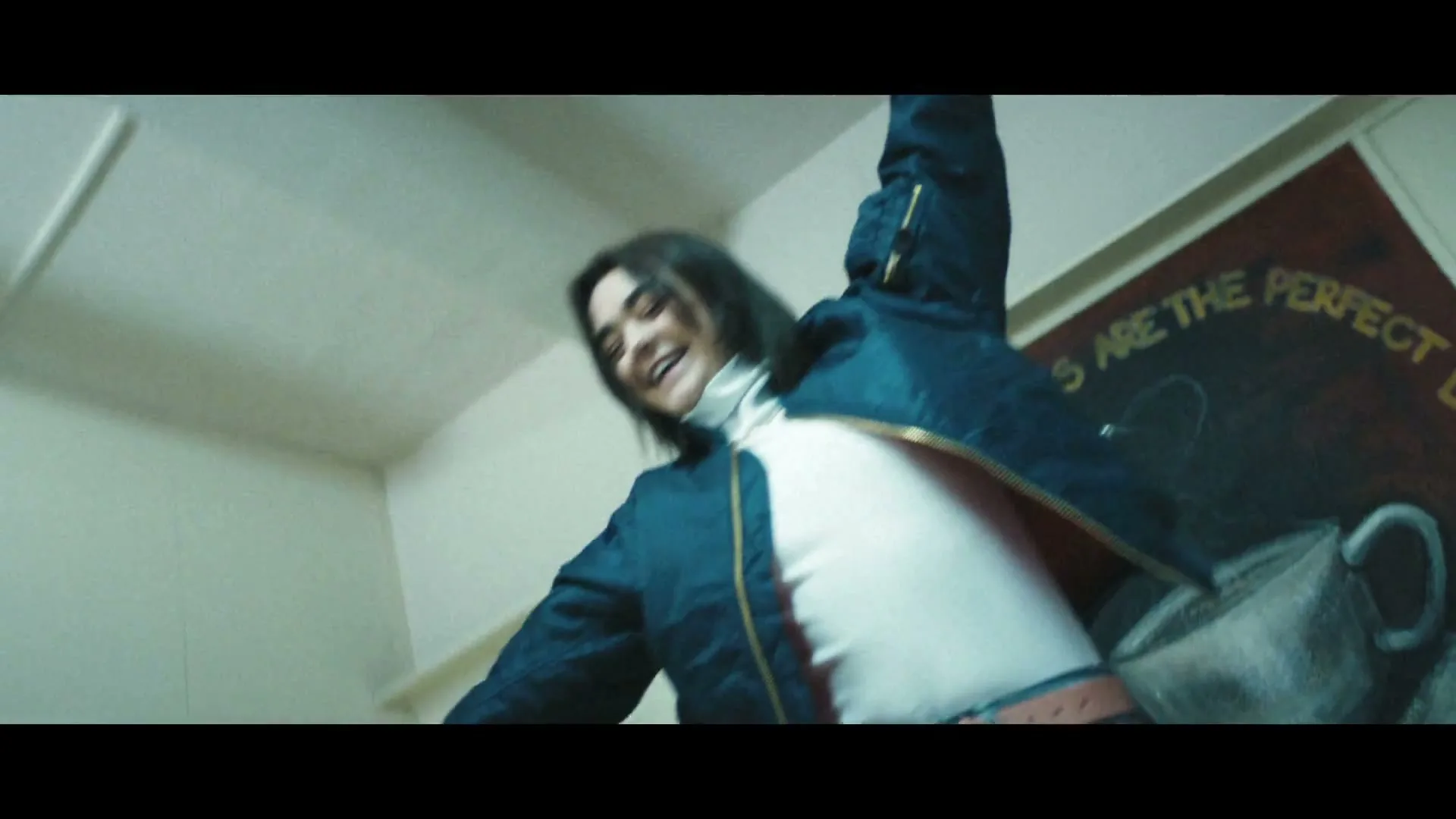 A young woman with dark hair, wearing a blue bomber jacket and jeans, dances joyfully in an interior room with a chalkboard sign.