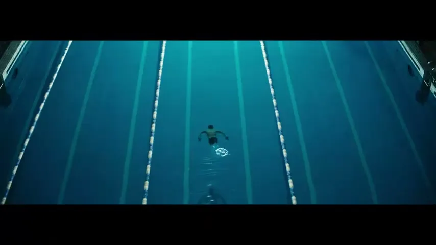 A male swimmer with a disability is seen from an overhead top-down angle, swimming in a blue pool with lane lines.