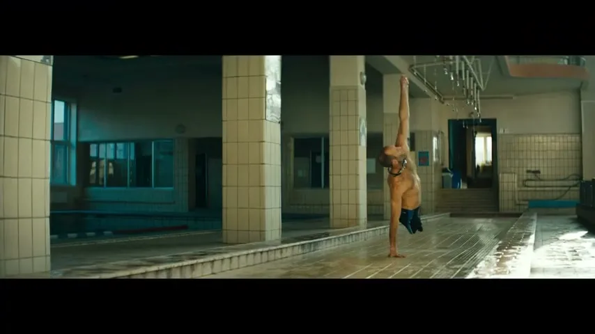 A male swimmer with an amputated leg performs a one-handed handstand on the edge of a swimming pool in an old, tiled changing room.