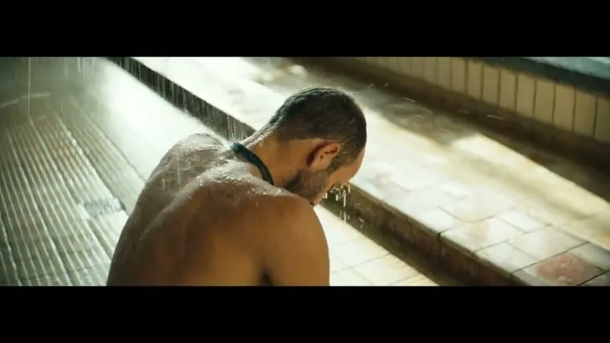 A bald man with a beard, wearing a black necklace, is seen from behind as he splashes water on his face in a swimming pool.