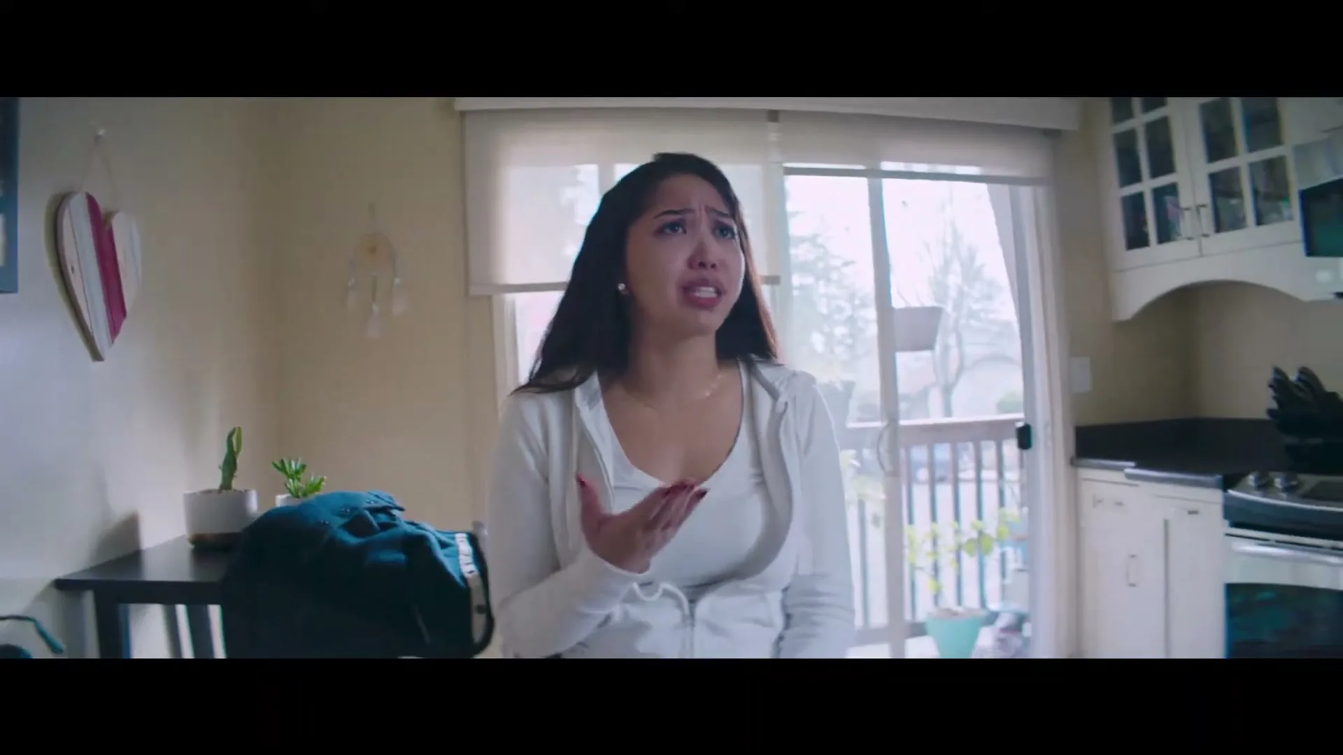 A young woman with long dark hair, wearing a white hoodie, stands in a kitchen, looking distressed and gesturing with her hands.
