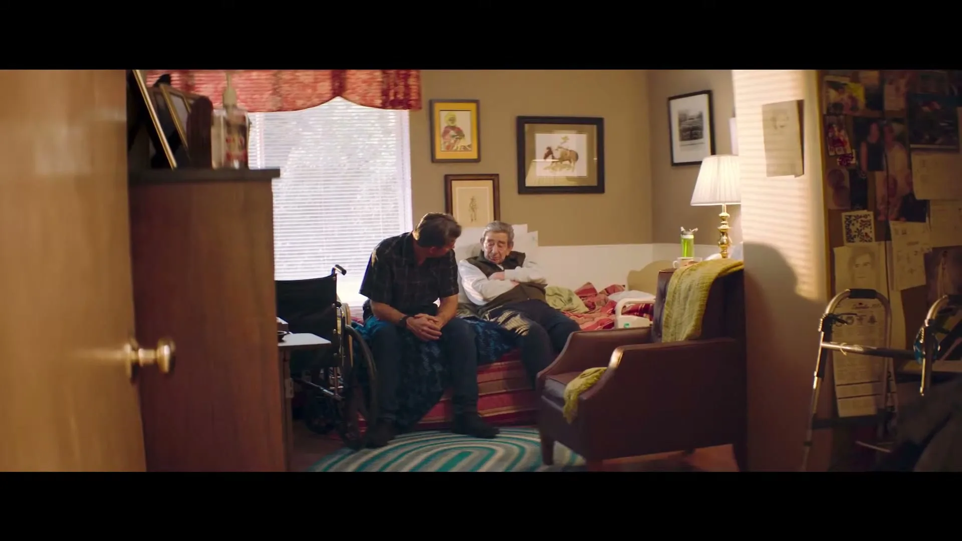 A middle-aged man sits on a bed next to an elderly man, both looking downcast in a sunlit bedroom.