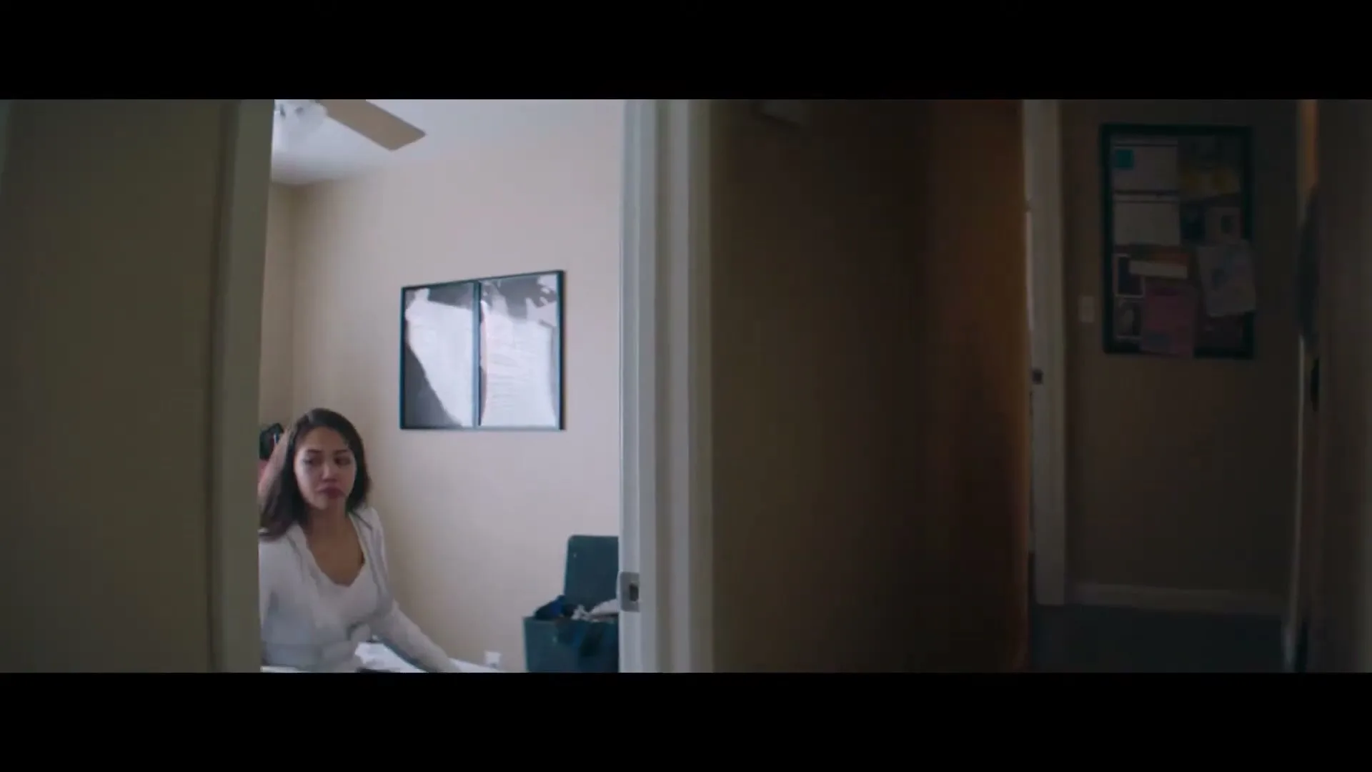 A young woman in a white shirt sits on a bed, looking concerned, as a man walks away down a hallway.