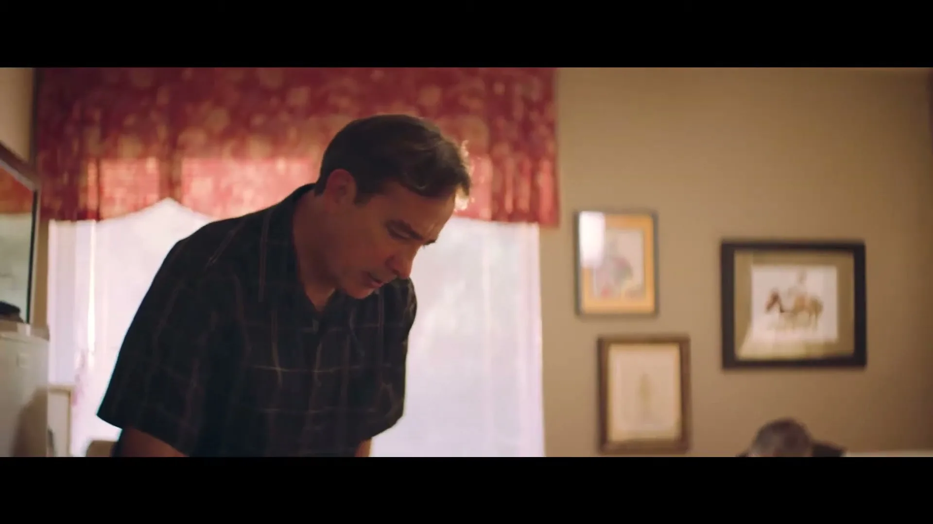 A middle-aged man in a plaid shirt looks down at something, while an older man is visible in the background, in a warm-toned room.