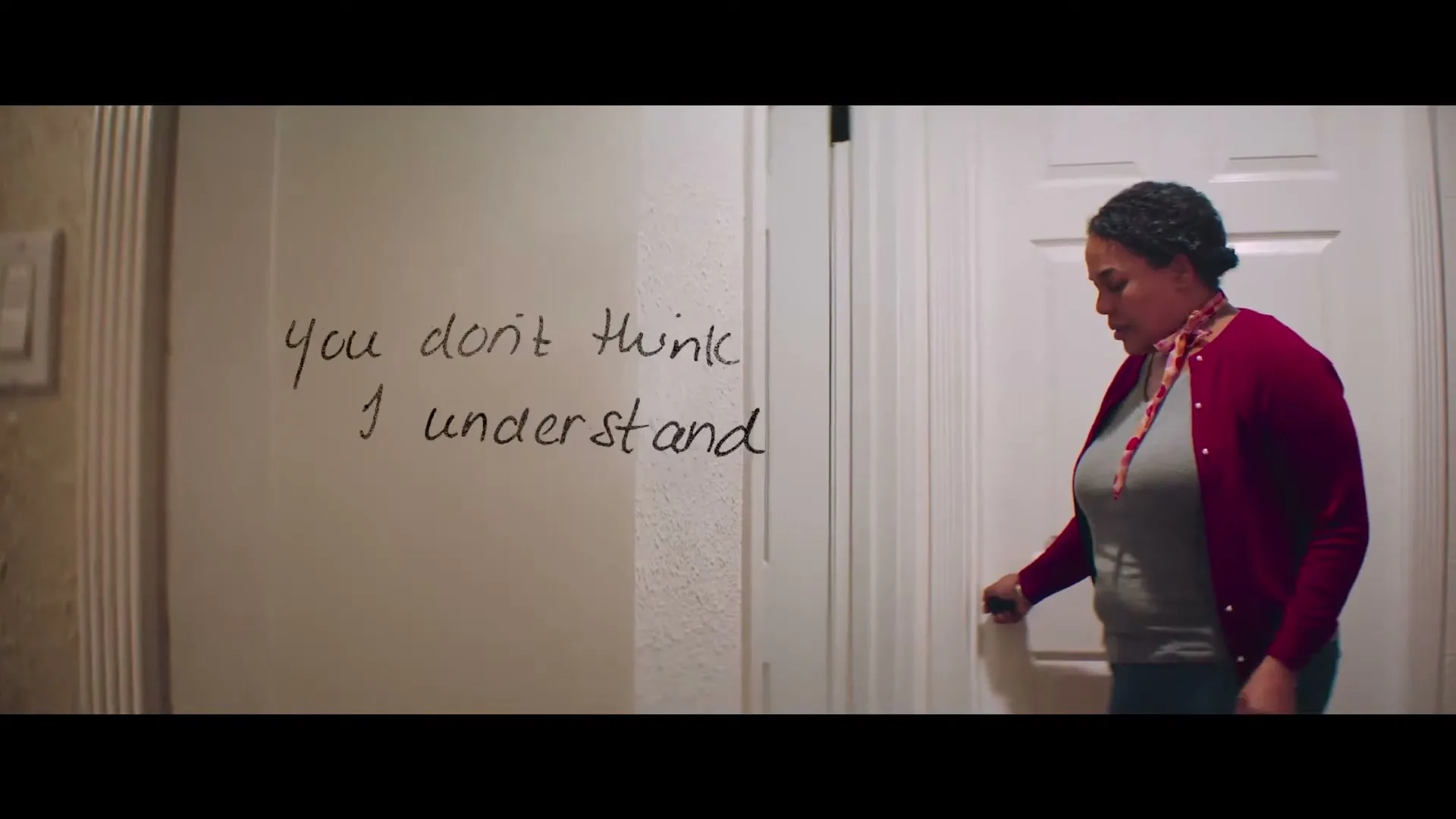 A woman with gray hair and a red cardigan opens a white door, revealing a message written on the wall next to it.